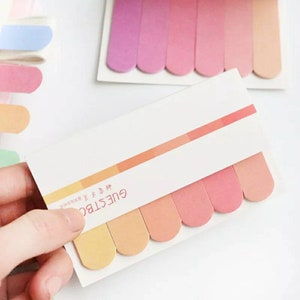 May include: A pack of colorful index tabs with rounded edges. The tabs are in shades of pink, orange, and yellow. The pack has the text "GUESTBOOK" printed on it.