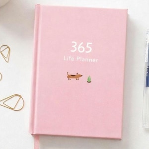 May include: A pink hardback planner with the text "365 Life Planner" in white. A small illustration of a dachshund and a tree are below the text. Gold paperclips and a pen are visible in the background.