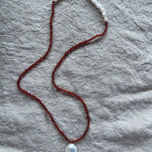 Op de afbeelding: Een ketting met een grote, iriserende witte barokke parel hanger. De ketting heeft een streng van kleine, roodbruine kralen en witte parels bij de sluiting. De sluiting is zilverkleurig.