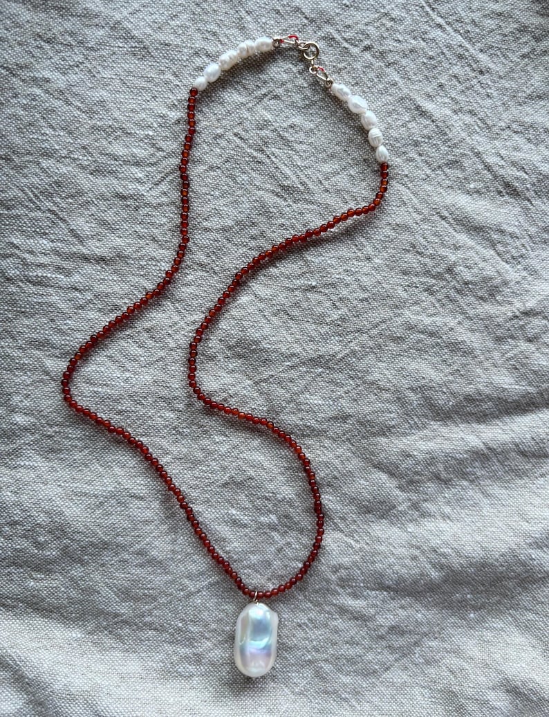 Op de afbeelding: Een ketting met een grote, iriserende witte barokke parel hanger. De ketting heeft een streng van kleine, roodbruine kralen en witte parels bij de sluiting. De sluiting is zilverkleurig.