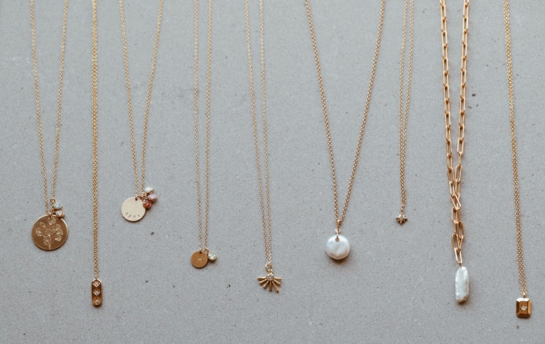 Puede incluir: Una colecci&oacute;n de diez collares de oro con diferentes colgantes, incluyendo una flor, una estrella, una perla, un c&iacute;rculo y un cuadrado. Los collares se muestran sobre un fondo gris claro.
