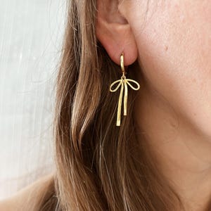 May include: Gold bow-shaped dangle earrings with a hoop design. The earrings are made of a textured metal.