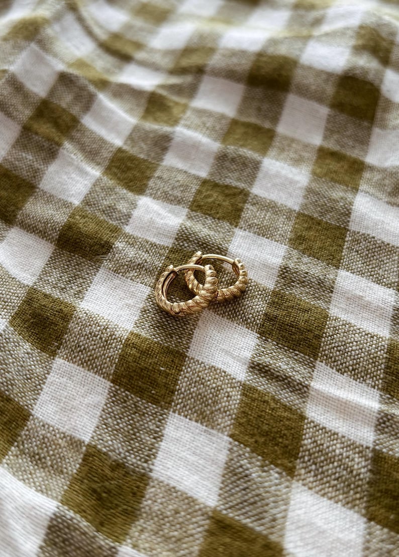 K&ouml;nnte beinhalten: Ein Paar goldene Creolen mit einem gedrehten Design. Die Ohrringe liegen auf einem wei&szlig;en und gr&uuml;nen Gingham-Hintergrund.