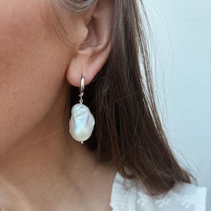 May include: A single, large, white baroque pearl earring with a silver hoop. The pearl has a lustrous, iridescent sheen. The earring is shown on a person's ear, with the focus on the jewelry.