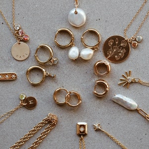 Puede incluir: Una colecci&oacute;n de collares y pendientes de oro con varios colgantes, incluyendo una perla, una flor, una estrella y una barra con tres peque&ntilde;os diamantes. Algunos de los colgantes tienen peque&ntilde;as piedras preciosas.
