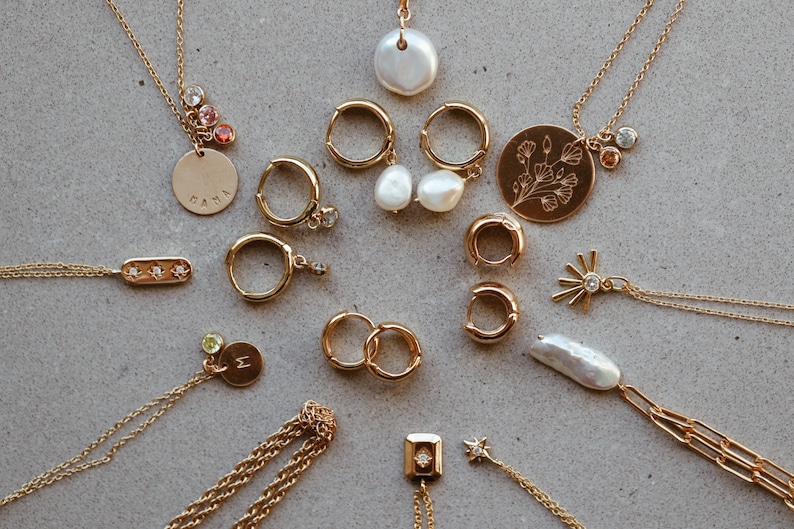 Puede incluir: Una colecci&oacute;n de collares y pendientes de oro con varios colgantes, incluyendo una perla, una flor, una estrella y una barra con tres peque&ntilde;os diamantes. Algunos de los colgantes tienen peque&ntilde;as piedras preciosas.