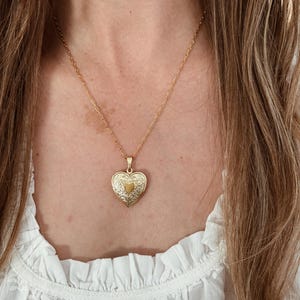 May include: A gold heart-shaped locket necklace. The locket features an ornate design and hangs from a delicate chain. The necklace is worn against a white blouse.