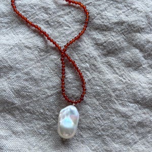 Op de afbeelding: Een ketting met een streng kleine, roodbruine kralen. Het heeft een grote, onregelmatig gevormde, witte parel hanger met iriserende highlights. De ketting wordt gepresenteerd op een getextureerd, lichtgrijs oppervlak.