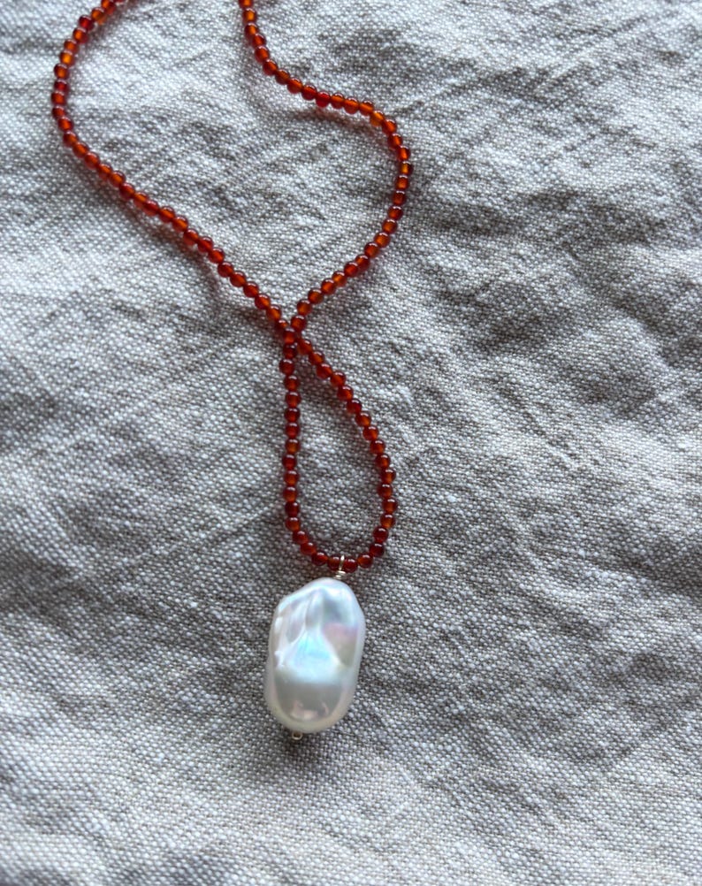 Op de afbeelding: Een ketting met een streng kleine, roodbruine kralen. Het heeft een grote, onregelmatig gevormde, witte parel hanger met iriserende highlights. De ketting wordt gepresenteerd op een getextureerd, lichtgrijs oppervlak.