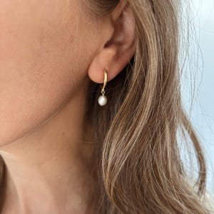 May include: A gold hoop earring with a small, white pearl pendant. The earring is worn in the ear of a person with blonde hair. The earring is a simple, elegant design.