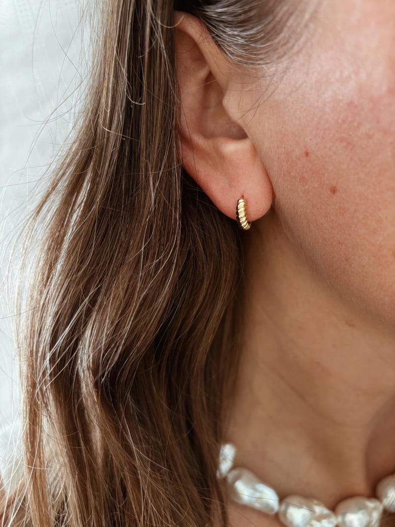 K&ouml;nnte beinhalten: Goldene Creolen mit einem strukturierten, perligen Design. Die Ohrringe werden am Ohr einer Person getragen.