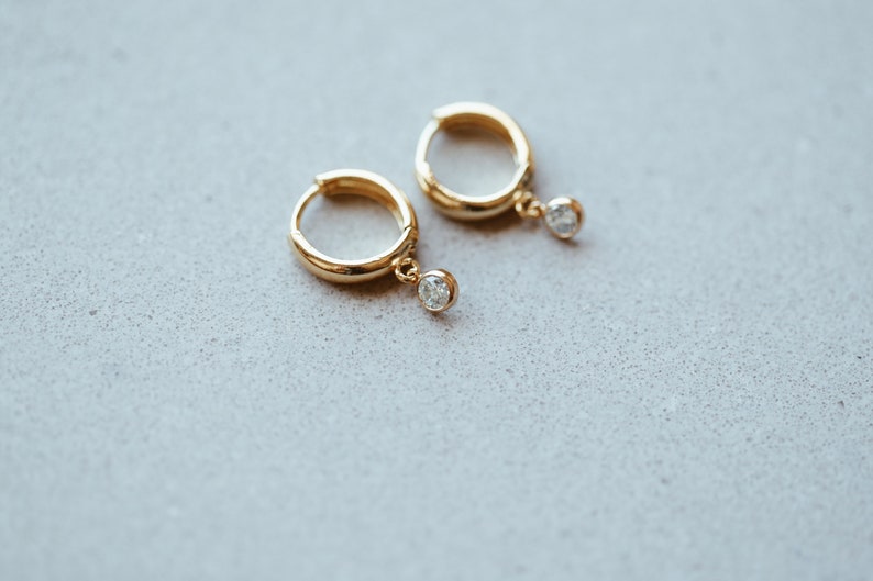 May include: A pair of gold hoop earrings with a small clear stone dangling from each hoop.