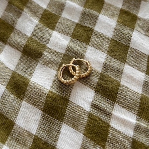 K&ouml;nnte beinhalten: Ein Paar goldene Creolen mit einem gedrehten Design. Die Ohrringe liegen auf einem wei&szlig;en und gr&uuml;nen Gingham-Hintergrund.