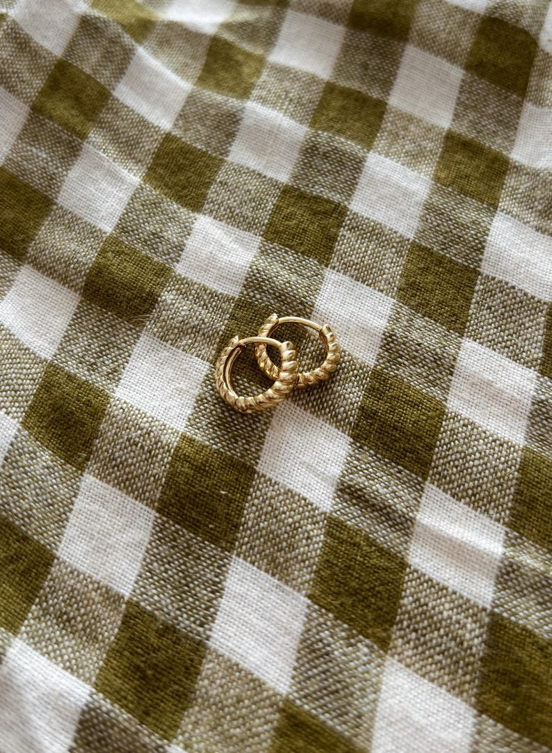 K&ouml;nnte beinhalten: Ein Paar goldene Creolen mit einem gedrehten Design. Die Ohrringe liegen auf einem wei&szlig;en und gr&uuml;nen Gingham-Hintergrund.