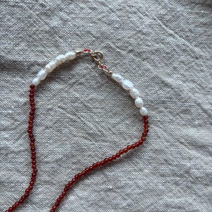 Op de afbeelding: Een delicate ketting met een streng kleine, ronde, rode kralen die overgaat in een streng witte, onregelmatig gevormde parels. De ketting is vastgemaakt met een goudkleurige sluiting.