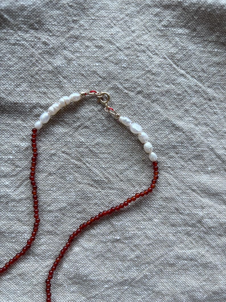 Op de afbeelding: Een delicate ketting met een streng kleine, ronde, rode kralen die overgaat in een streng witte, onregelmatig gevormde parels. De ketting is vastgemaakt met een goudkleurige sluiting.