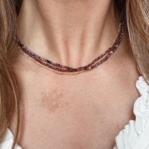 Può includere: Una collana a più fili con piccole perline colorate. La collana ha fili di perline bordeaux, viola e oro. La collana è indossata intorno al collo su uno sfondo bianco.