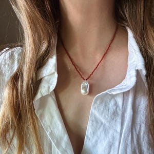 Op de afbeelding: Een delicate ketting met een grote, onregelmatige witte parel hanger, hangend aan een rode kralenketting. De ketting wordt gedragen over een wit overhemd.