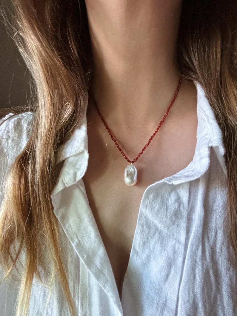 Op de afbeelding: Een delicate ketting met een grote, onregelmatige witte parel hanger, hangend aan een rode kralenketting. De ketting wordt gedragen over een wit overhemd.