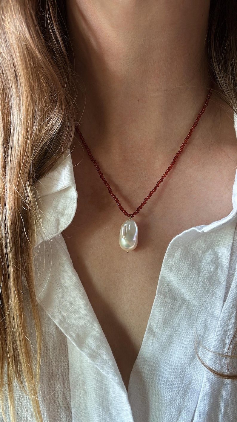 Op de afbeelding: Een ketting met een grote, onregelmatig gevormde witte parel hanger. De parel hangt aan een delicate streng van kleine, roodbruine kralen. De ketting wordt getoond tegen een wit overhemd.