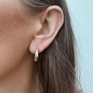 May include: A close-up of a person's ear wearing a gold teardrop-shaped earring. The earring is smooth and reflective, catching the light. The earring is a simple, elegant design.