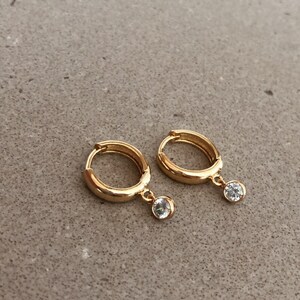 May include: A pair of gold hoop earrings with a small clear stone dangling from each hoop.