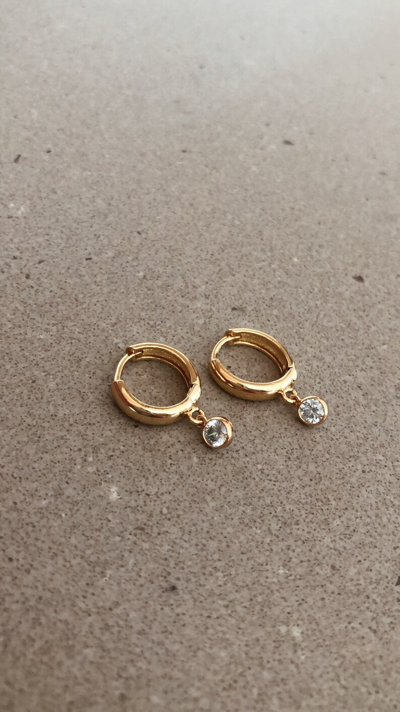 May include: A pair of gold hoop earrings with a small clear stone dangling from each hoop.