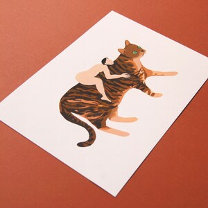 May include: A brown and black striped cat with green eyes is lying down. A nude person is sitting on the cat's back and hugging the cat's neck.