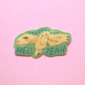 Puede incluir: Una pegatina con un diseño de búho de dibujos animados, con las alas extendidas y la frase "OHHHHHHH HELL YEAH" en azul. El búho es de color amarillo claro con detalles marrones, sobre un fondo verde. La pegatina está sobre un fondo rosa.