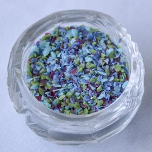 Puede incluir: Un tarro de vidrio transparente lleno de granas de colores. Las granas son una mezcla de azul, verde y rosa.
