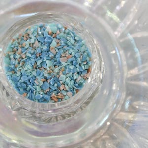 May include: A close-up of a clear glass bowl filled with small, blue and white iridescent glass pieces. The bowl is sitting on a white, textured surface.