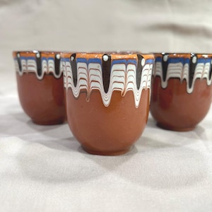 May include: Four brown ceramic cups with a decorative rim. The rim features a pattern of white, black, blue, and orange. The cups have a rounded shape and are arranged on a white surface.