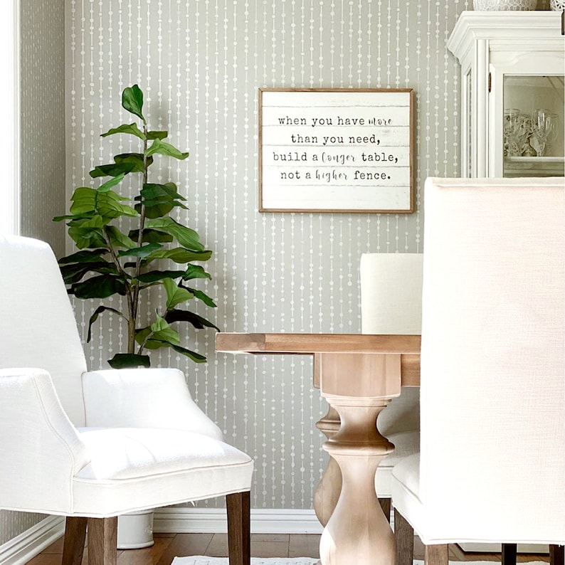 May include: A white dining room with a wooden table and white chairs. The wall is covered in a gray wallpaper with a white pattern. A framed sign with the text "when you have more than you need, build a longer table, not a higher fence." hangs on the wall.