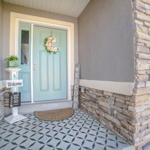 May include: A front porch with a light blue door, a welcome sign, and a floral wreath. The porch has a patterned floor and a stone accent wall. A small welcome sign is on a white stand.