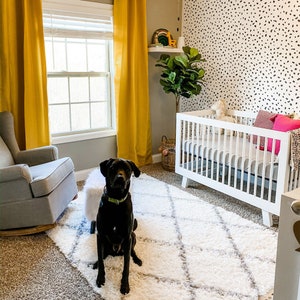 May include: A black Labrador Retriever dog sits on a white fluffy rug in front of a white crib with a black and white patterned wall behind it. The crib has a white mattress and a white blanket. The dog is looking at the camera.