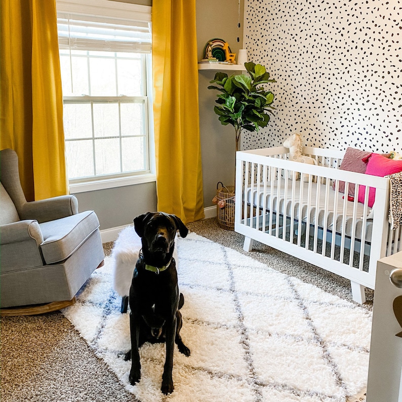 May include: A black Labrador Retriever dog sits on a white fluffy rug in front of a white crib with a black and white patterned wall behind it. The crib has a white mattress and a white blanket. The dog is looking at the camera.