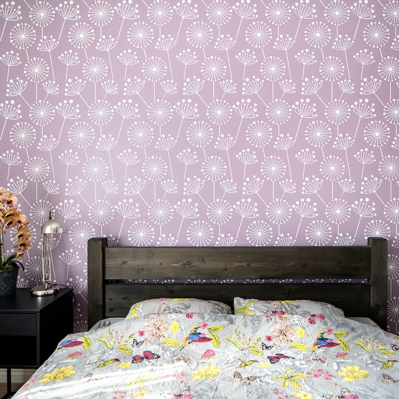 K&ouml;nnte beinhalten: Ein Schlafzimmer mit einem Bett mit einer Bettdecke und Kissen mit Blumenmuster. Das dunkle Holz-Kopfteil steht vor einer hellvioletten Wand, die mit einem wei&szlig;en Pusteblumenmuster verziert ist. Ein schwarzer Nachttisch mit einer Lampe und einer Topfpflanze.