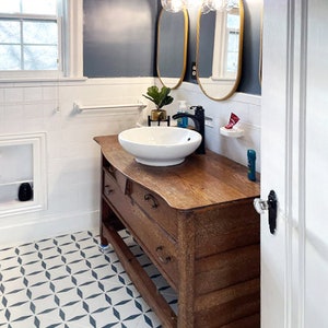 May include: Bathroom interior with a vintage wooden vanity, a white vessel sink, and a gold-framed mirror. The walls are painted a dark blue, and the floor features a geometric tile pattern. A modern light fixture with globe bulbs illuminates the space.