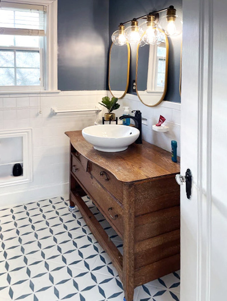 May include: Bathroom interior with a vintage wooden vanity, a white vessel sink, and a gold-framed mirror. The walls are painted a dark blue, and the floor features a geometric tile pattern. A modern light fixture with globe bulbs illuminates the space.