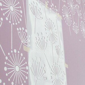 K&ouml;nnte beinhalten: Eine transparente Schablone mit einem Pusteblumen-Design ist an einer hellvioletten Wand befestigt. Die Wand zeigt ein sich wiederholendes Muster aus wei&szlig;en Pusteblumen-Silhouetten. Die Schablone wird verwendet, um das gleiche Design zu malen.
