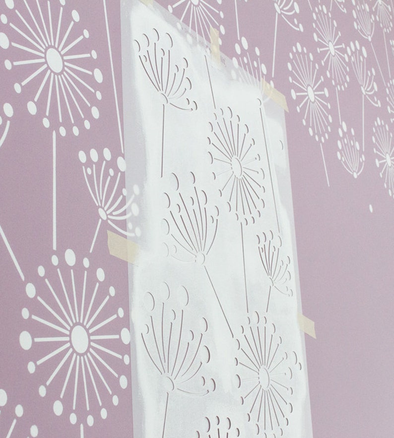 K&ouml;nnte beinhalten: Eine transparente Schablone mit einem Pusteblumen-Design ist an einer hellvioletten Wand befestigt. Die Wand zeigt ein sich wiederholendes Muster aus wei&szlig;en Pusteblumen-Silhouetten. Die Schablone wird verwendet, um das gleiche Design zu malen.