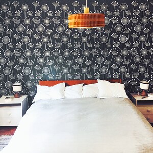 K&ouml;nnte beinhalten: Schlafzimmer mit Bett, Nachttischen und gemusterter Tapete. Das Bett hat wei&szlig;e Kissen und eine graue Bettdecke. Die Wand ist dunkel mit einem wei&szlig;en Blumenmuster. &Uuml;ber dem Bett h&auml;ngt eine gewebte Pendelleuchte.