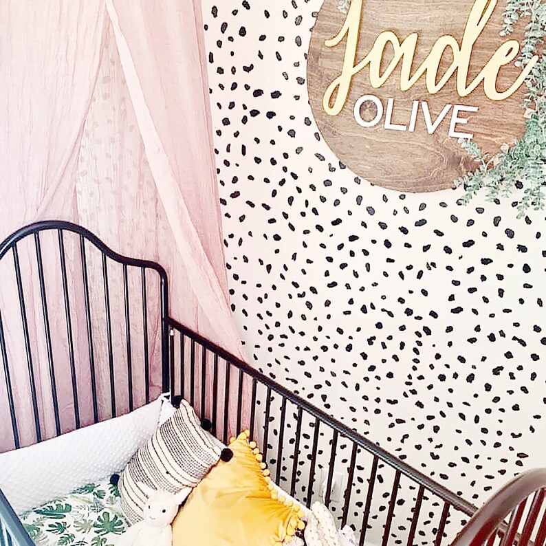 May include: A black metal crib with a white and green patterned sheet and a yellow pillow. The crib is in a room with a pink curtain and a white wall with black polka dots. A wooden sign with the name "Jade Olive" is hanging on the wall.