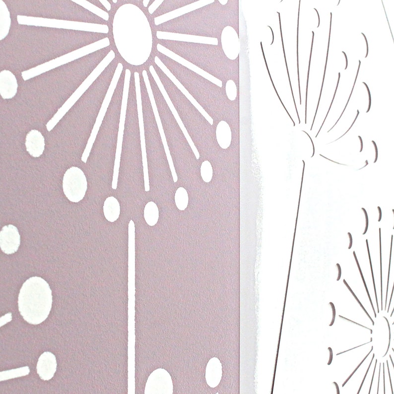 K&ouml;nnte beinhalten: Nahaufnahme einer dekorativen Platte mit einem Pusteblumen-Design. Die Platte hat einen mauvefarbenen Hintergrund mit wei&szlig;en, ausgeschnittenen Pusteblumenformen. Das Design umfasst einen zentralen Kreis mit radialen Linien und kleineren Kreisen.