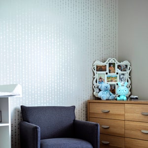 May include: A light blue armchair sits in front of a wall with a white beaded pattern. A dresser with wooden drawers is to the right of the chair. A white picture frame with multiple photos is on top of the dresser. Two blue stuffed animals are in front of the frame.