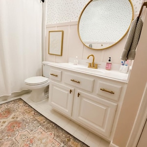 May include: A white bathroom vanity with gold hardware and a white countertop. The vanity has three drawers and two doors. A round gold mirror hangs above the vanity. A white toilet is visible in the background.