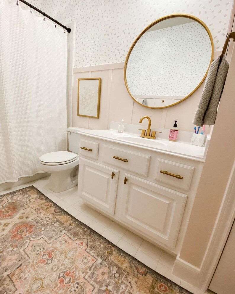 May include: A white bathroom vanity with gold hardware and a white countertop. The vanity has three drawers and two doors. A round gold mirror hangs above the vanity. A white toilet is visible in the background.
