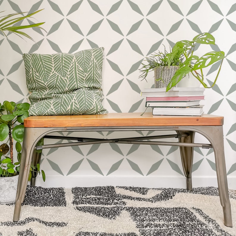 May include: A wooden bench with metal legs supports a green leaf-patterned pillow, books, and potted plants. The bench is set against a white wall with a gray diamond pattern. A patterned rug is on the floor.