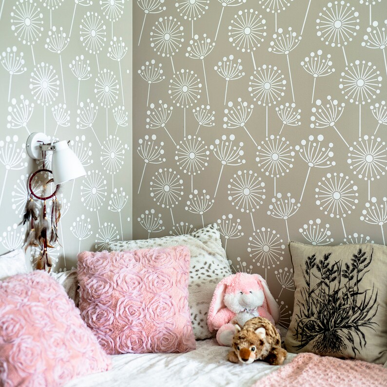K&ouml;nnte beinhalten: Eine Zimmerecke mit beigefarbenen W&auml;nden, die mit einem wei&szlig;en Pusteblumenmuster verziert sind. Rosa Kissen mit Rosenstruktur, ein Pl&uuml;schhase und ein dekoratives Kissen mit schwarzem Blumendesign liegen auf einem wei&szlig;en Bett. Eine wei&szlig;e Lampe und ein Traumf&auml;nger sind an der Wand befestigt.