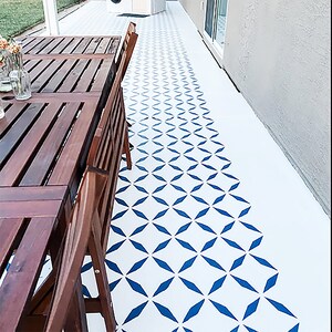May include: Outdoor patio with a white floor featuring a blue geometric pattern. A dark brown wooden table and chairs are on the left. The floor pattern transitions from solid blue shapes to a dotted design.